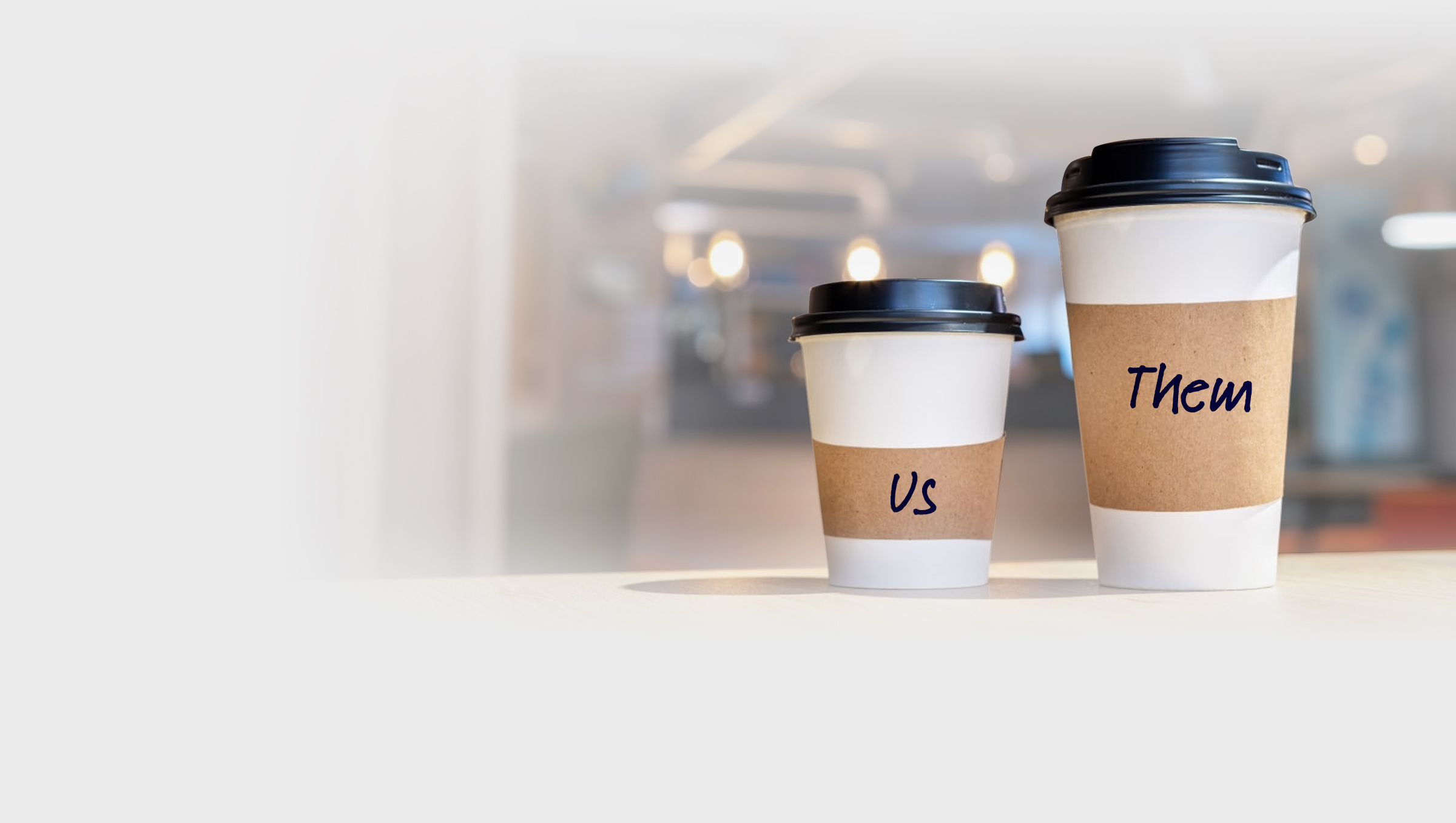 Small and large paper coffee cups on a desk in an office. 'Us' written in marker on the small cup 'Them' written on the large cup